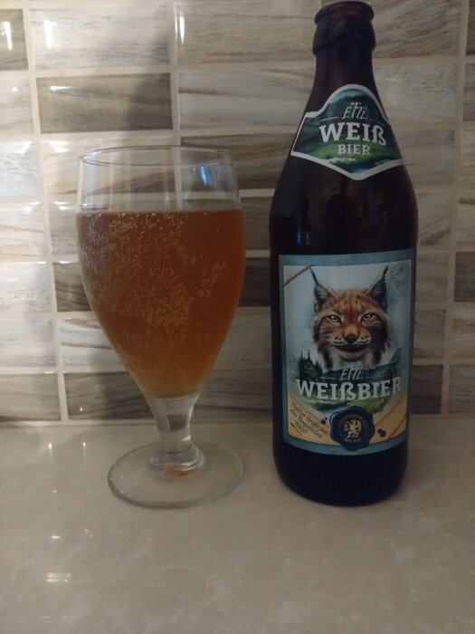 En flaska Ettaler Weissbier och ett glas öl på en ljus bänkskiva med tegelmönstrad vägg i bakgrunden.