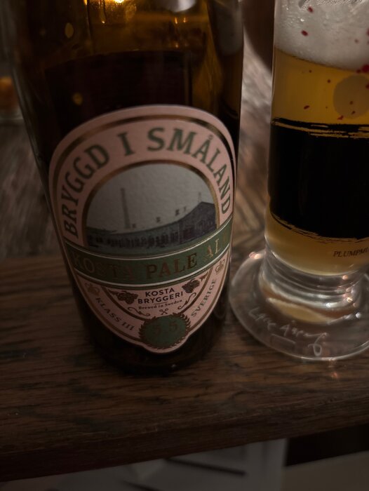 Flaska med etikett "Kosta Pale Ale, Bryggd i Småland" och ett glas öl med skum; fotograferade på träbord.