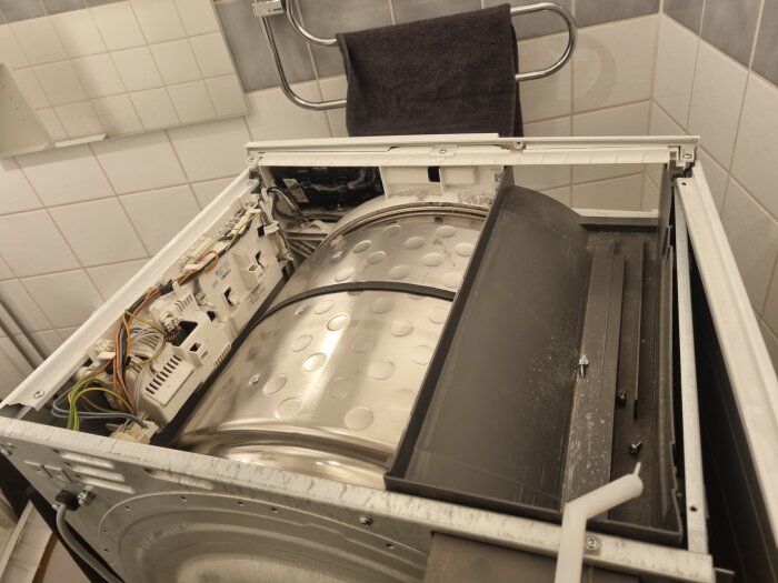 Öppen torktumlare med synlig metalltrumma och elektriska komponenter, fotad ovanifrån i ett badrum.