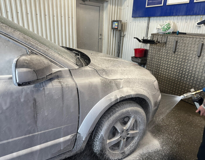 Bil i tvätthall täckt med skum från en högtryckstvätt, med fokus på främre delen och hjulhus, person som tvättar med lans.