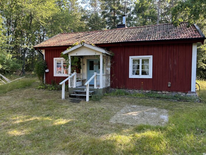 Rött trähus med tegeltak, klassisk torpargrund, vita fönster och entrétrappa, omgiven av gräs och skog.