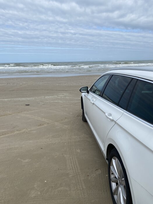 Vit bil parkerad på en sandstrand med vågor i bakgrunden under en molnig himmel.
