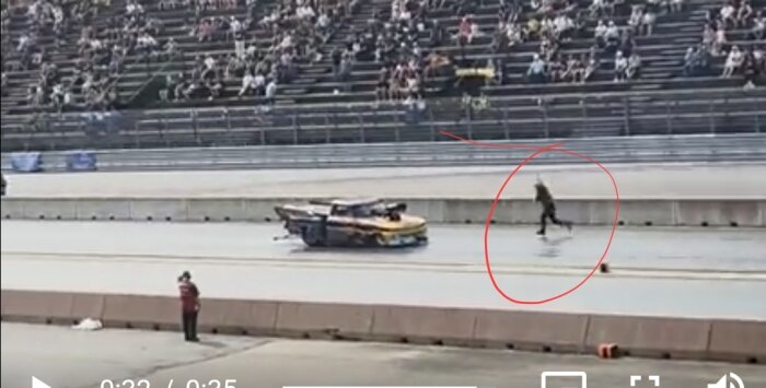 Person springer över en racerbana medan en bil backar, publik i bakgrunden.