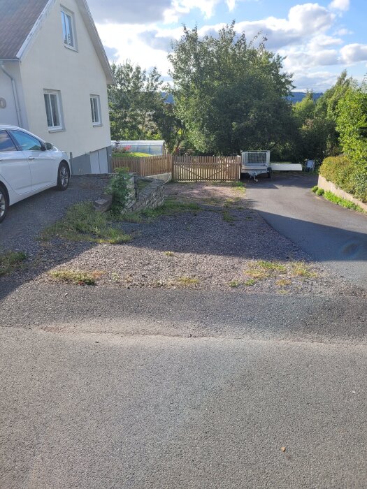 Uppfart med grus och asfalt, omgiven av träd och byggnader; gamla sliprar som stöd ska ersättas med L-stöd.