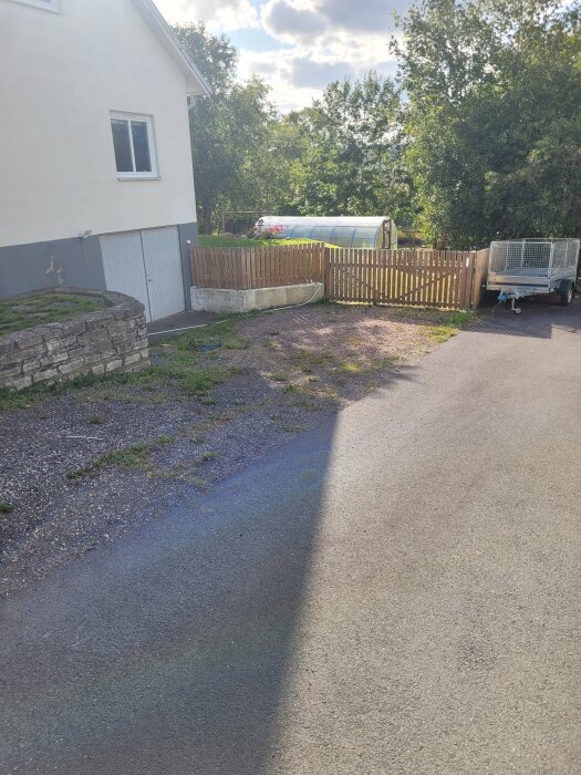 Nedfart och parkering vid hus, omgiven av gamla slipers och grus, bredvid en vägg och ett trästaket med släp och växthus i bakgrunden.