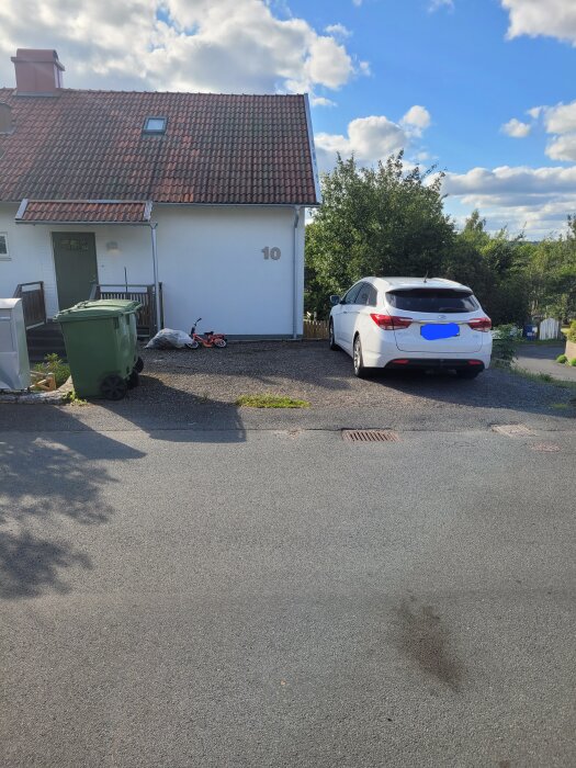 Parkering med en vit bil och gamla slipers som avgränsar, en liten cykel och soptunnor syns vid sidan av ett vitt hus med rött tak.