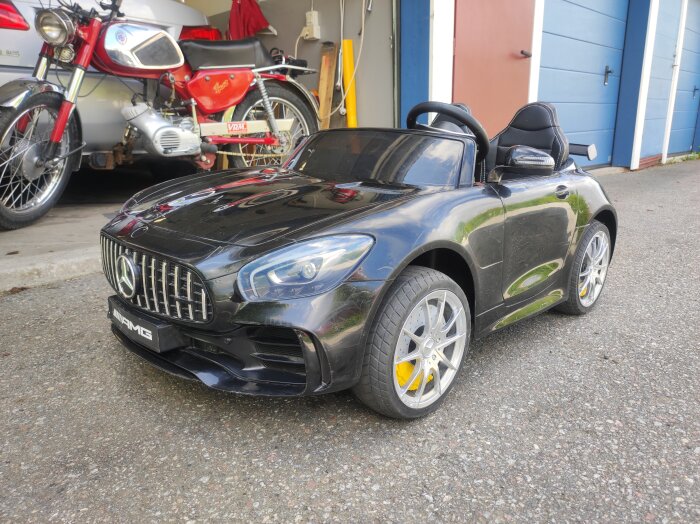 Leksaksbil i form av en svart Mercedes AMG, parkerad bredvid en röd motorcykel i ett garageområde.