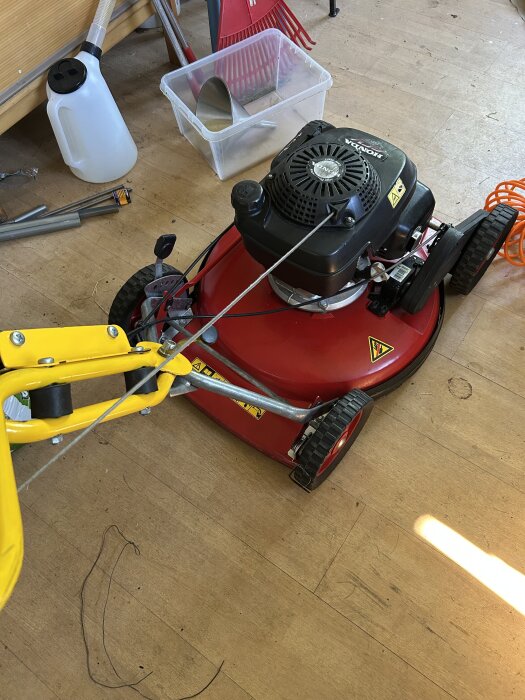 Röd gräsklippare med gul handtag står på trägolv, omgiven av verktyg och trädgårdsredskap.