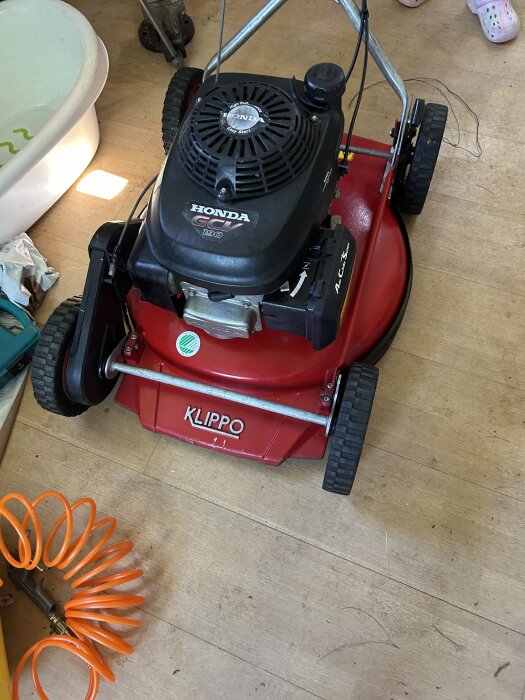 Röd Klippo gräsklippare med Honda motor, placerad inomhus på ett trägolv med en orange luftslang nära.