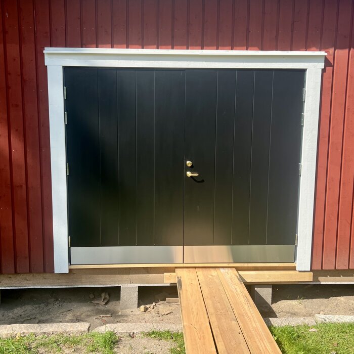 Svart dubbeldörr med vita karmar på röd träfasad, upphöjd på plintar, saknar golv.