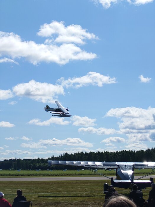 Ett sjöflygplan flyger över ett fält mot en klarblå himmel med moln, med ett annat flygplan och åskådare synliga i förgrunden.