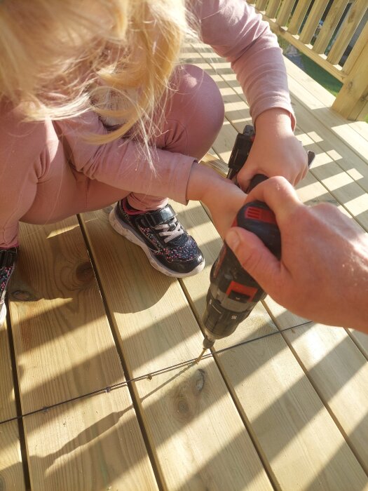 Barn och vuxen arbetar tillsammans med att skruva trallbrädor på en veranda i solen.