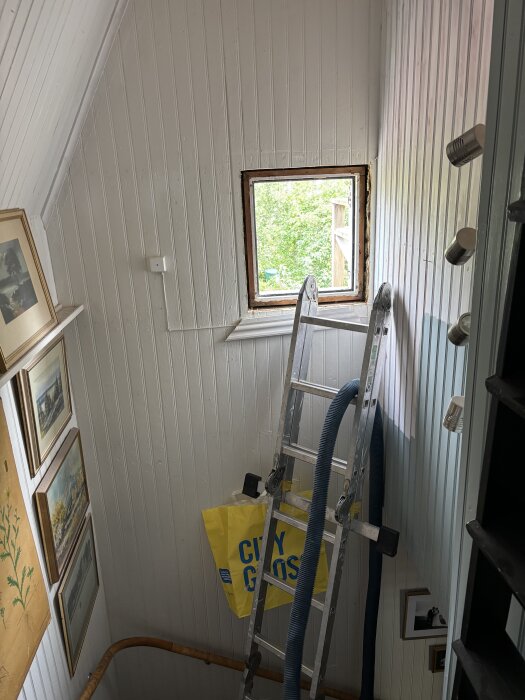 Stege vid nytt fönster på vit trävägg, lampa installerad ovanför, tavlor på väggen, gul City Gross-påse hänger på stegen.