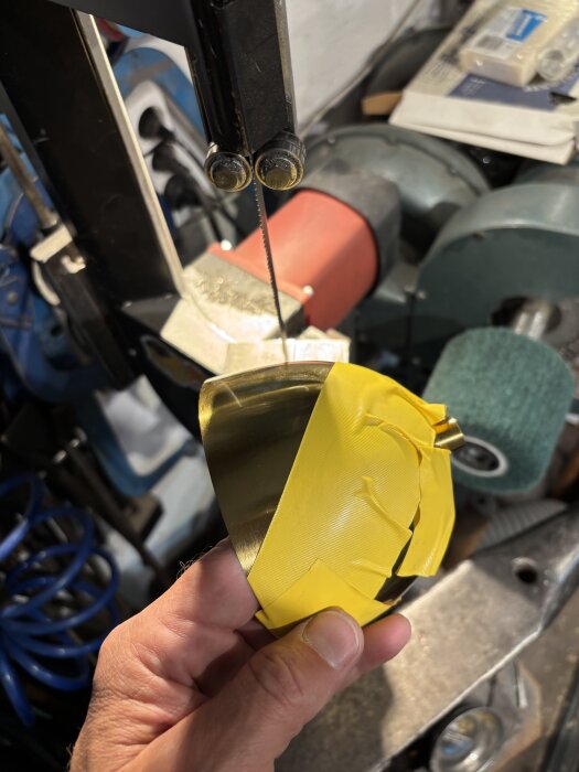 En hand håller en gul tejpad metallbit framför en bandsåg i en verkstad.