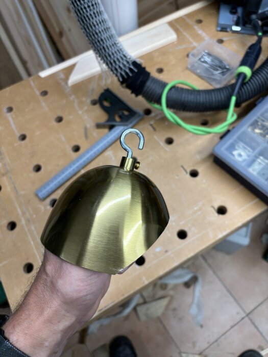 Man håller en metallkuppellampa i mässing över en verkstadsbänk med verktyg och tillbehör i bakgrunden.