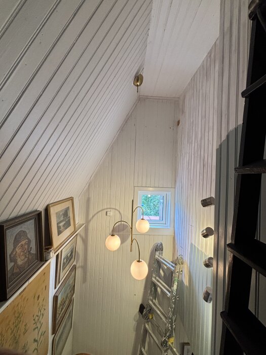 Nyinstallerad lampa med tre ljuskällor i trappuppgång. Nymålad vit träpanel, nytt fönster och dekorativa tavlor syns. Stege mot väggen.