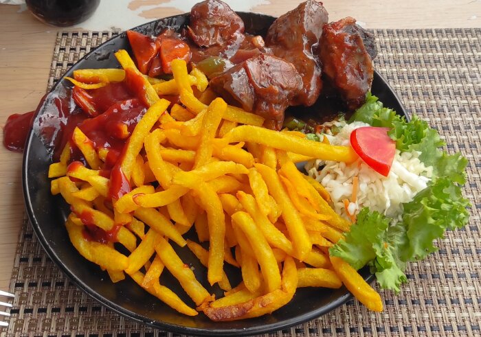 Svart tallrik med getkött utan ben, pommes frites med ketchup, sallad med tomat, och ris. Placering på bord vid restaurang nära Kampala bussstation.