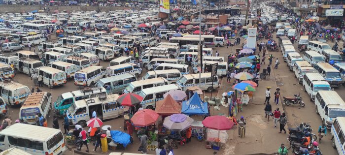 Bussstation i centrala Kampala full av vita minibussar och marknadsstånd täckta av färgglada parasoller med människor runt omkring.