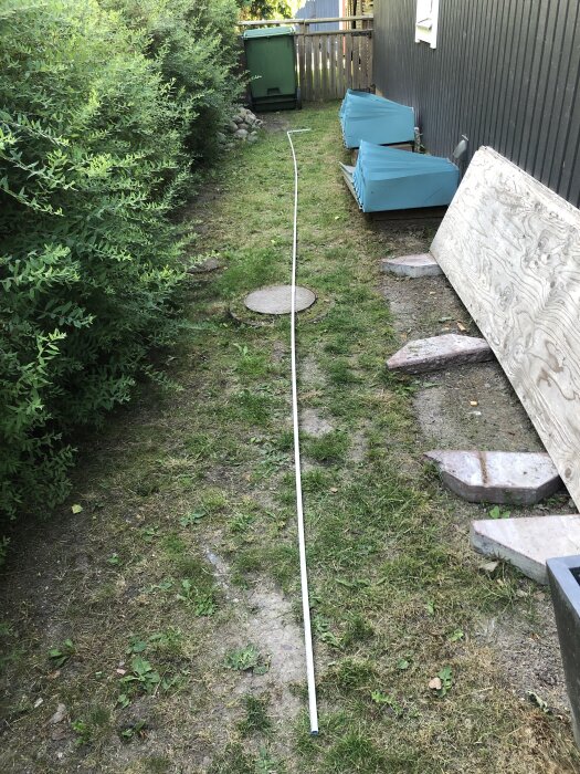 En trädgårdsgång med gräs och en vit pinne på marken, omgiven av buskar och byggmaterial längs en fasad.