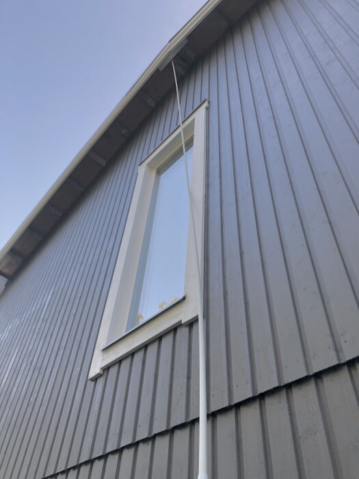 Grå fasad på ett hus med utstickande fönsterkarm och hängränna synlig, sedd underifrån mot en blå himmel.