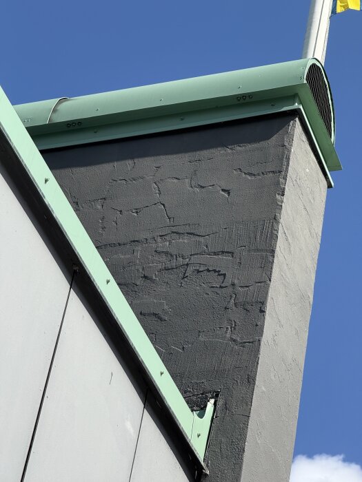 Skorsten med avflagad puts och övermålad yta, grå fasad med gröna detaljer mot blå himmel.