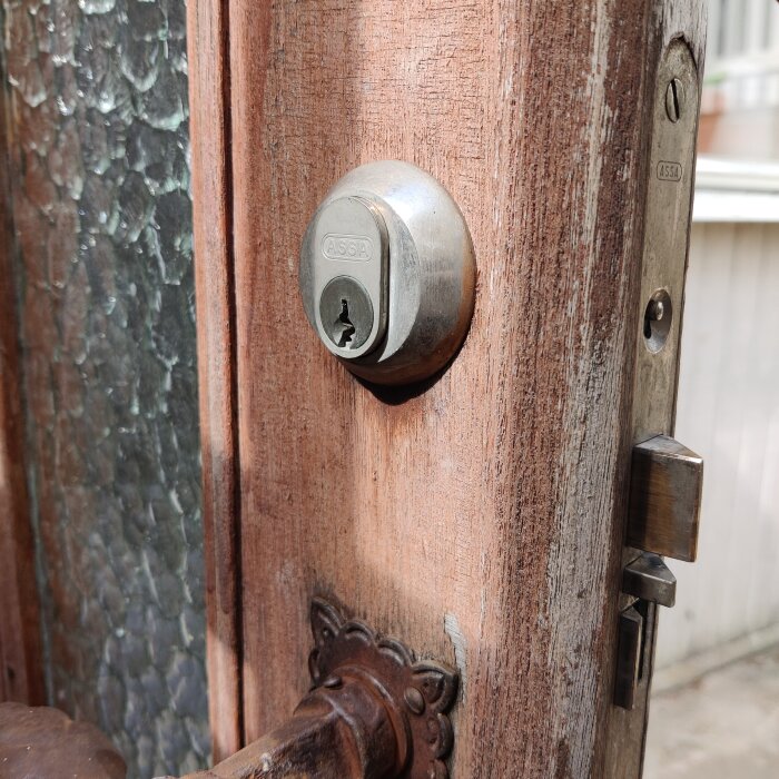 Gammalt entrélås med vred på insidan av en trädörr, bredvid en glasruta med textur och synligt låshus.