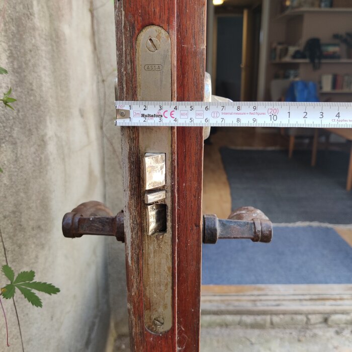 Gammalt dörrlås på trädörr med märket "ASSA," mätt med måttband. Insidan av ett rum syns i bakgrunden.