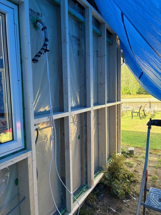 Väggkonstruktion under byggnation, med isolering och fönsterkarm, täckt av en blå presenning, och synlig trädgård i bakgrunden.