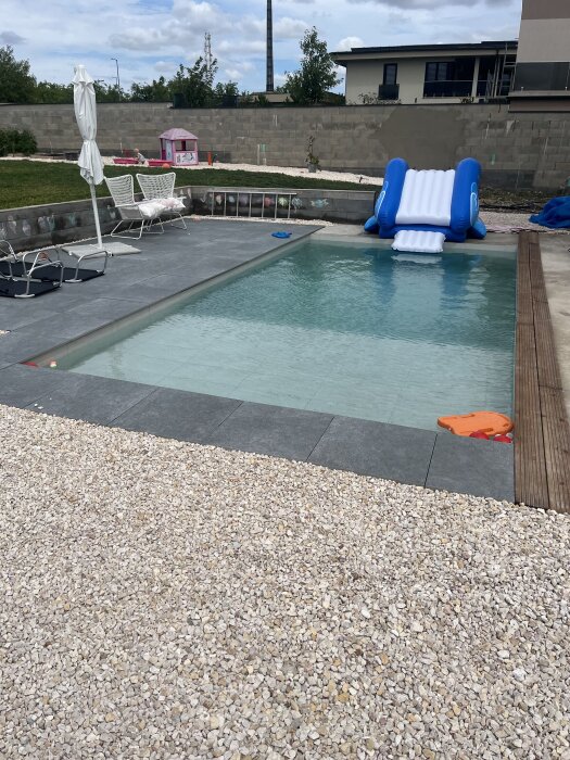 Poolområde med halvfärdig terrass, solsängar, paraply, blå uppblåsbar rutschkana och småbarnshus i bakgrunden.
