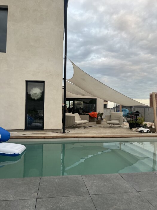 En halvfärdig terrass med solsängar och solsegel bredvid en pool och ett modernt hus.