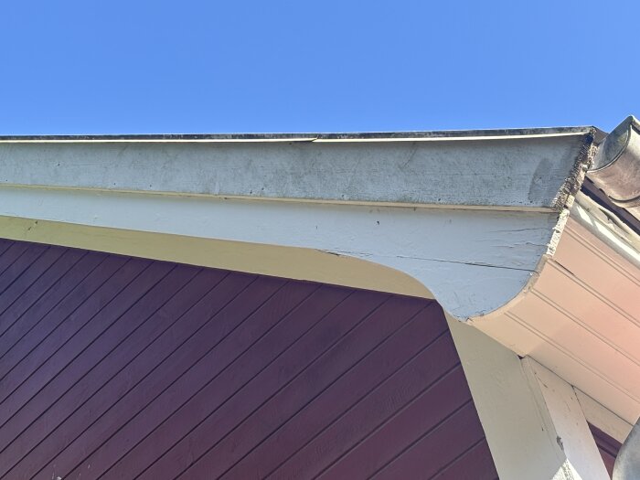 Hörn av ett hus takfot med vit och röd träpanel, skadad överliggare, tydligt synligt vid en klar blå himmel.