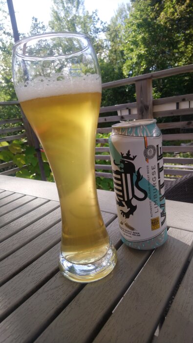 Belgisk witbier i ett högt glas på ett bord bredvid en burk med etikett i solen, omgiven av grönska i bakgrunden.