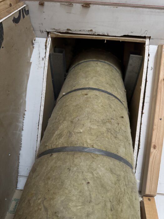 Isolerat rökrör genom en genomföring i taket med träpaneler och brännbart material synligt på sidorna.