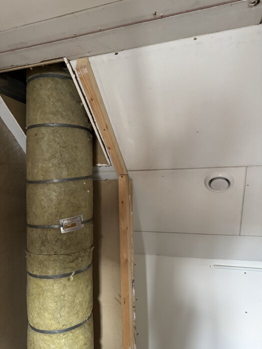 Isolerat rökrör i genomföring på vinden, omgiven av isolering och träpanel. Frånluftsventil synlig i väggen.