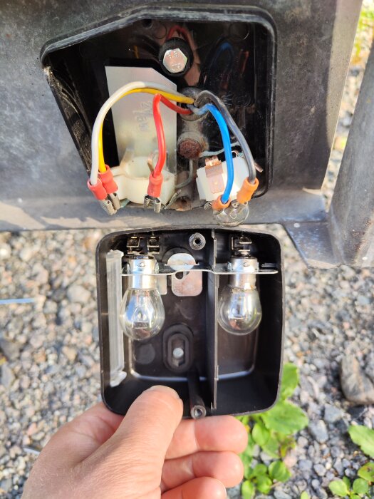 Baklampa på släp med två glödlampor och flera färgade elkablar visas; handen håller i lamphusets lock.