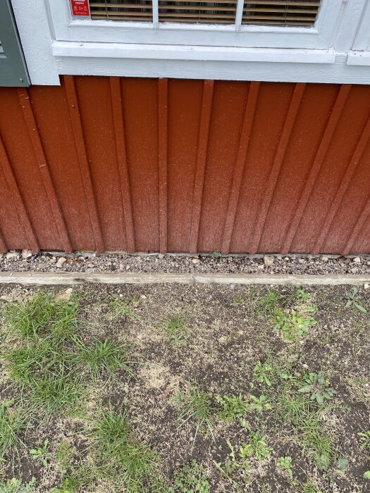 Nymålad röd vägg med vitt fönster, smutsstänk på nederdelen. Marken framför har gles grästillväxt och stenar närmast väggen.