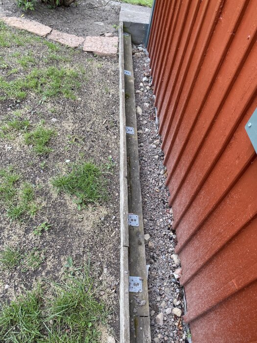 Röd vägg med grus och stenar vid basen, gräs och jord intill, indikerande potentiella orsaker till smutsstänk på nymålad yta.