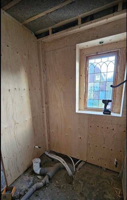 Väggar klädda med plywood i ett rum under konstruktion, med synliga rör och ett fönster med spröjs. En skruvdragare står på fönsterbrädan.