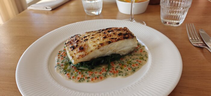 Grillad fisk på en bädd av spenat med krämig sås och röd kaviar, serverad på en vit tallrik vid bordet.