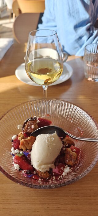Dessert med bär och glass serverad på glasfat, bredvid ett glas vitt vin på ett träbord vid middag på Tylösand strand.