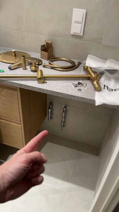 Guldiga rördelar och kopplingar på en bänkskiva i ett badrum under installation, med ett finger pekande mot rören under bänken.