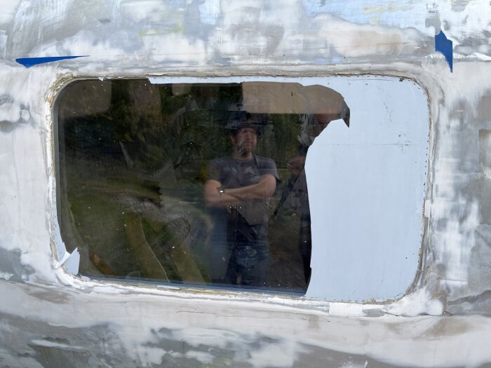 Ett fönster på en bastuvagn med sprucken skyddsfilm och reflektion av en person i rutans yta.