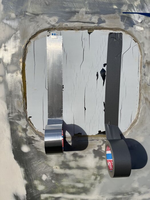 Två typer av tejp applicerade på en sprucken skyddsfilm på ett fönster, omgiven av en ojämn, målad yta.