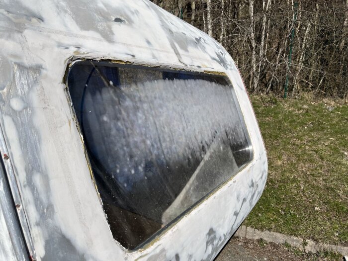 Fönster med sprucken skyddsfilm på en bastuvagn, visar tecken på väderpåverkan och fuktläckage, omgiven av ett skogsområde och grönska.