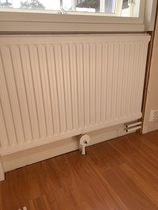 Vitt element under fönster med nya kopparrör som sticker upp genom golvet och ansluter till elementet. Golv av trä och eluttag till höger.