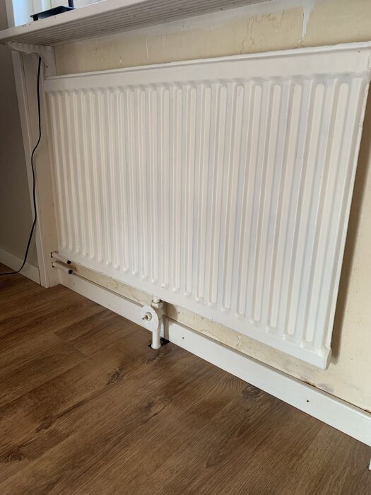 Vitt element med nya kopparrör anslutna, rören går igenom golvet och ansluter till botten av elementet. Golv med träimitation i ett rum i ett hus.