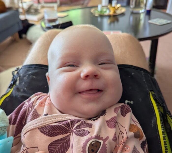 En bebis i blommig klädsel sitter i knäet på en person med arbetsshorts, med ett bord i bakgrunden.