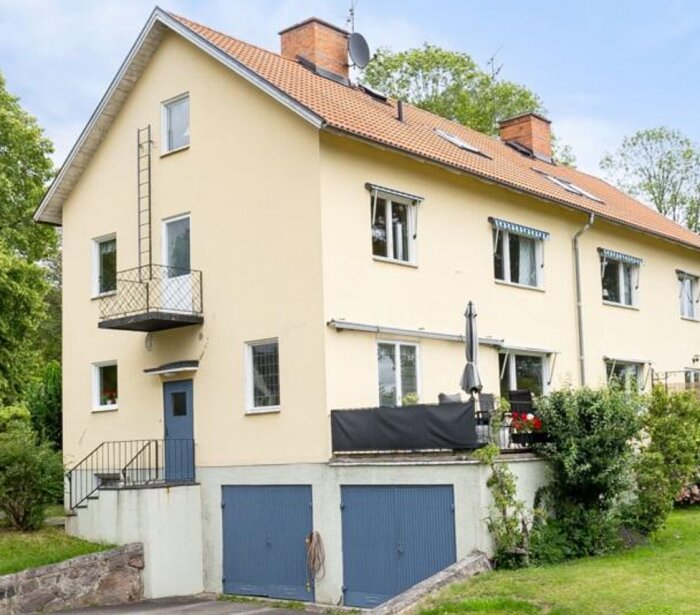 Utomhusbild av ett beige flerfamiljshus med blå dörrar och garage, en liten balkong och en altan med parasoll och svarta detaljer.