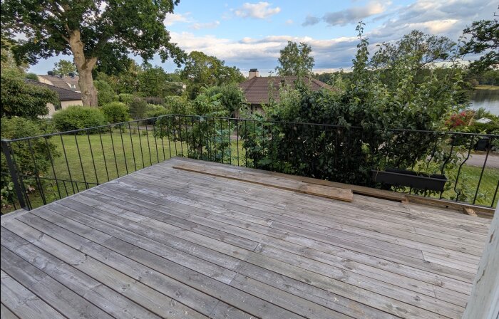 Trädäck med träplankor, inramat av ett gammalt gjutjärnsräcke, omgiven av grönskande träd och buskar, med utsikt över trädgård och hus.