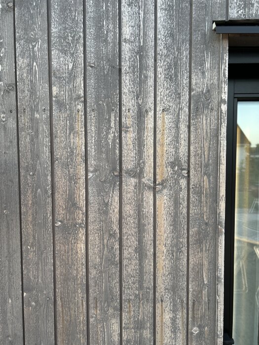 Träpanelfasad med flagnande färg efter borstning, vid ett fönster med solreflektion.